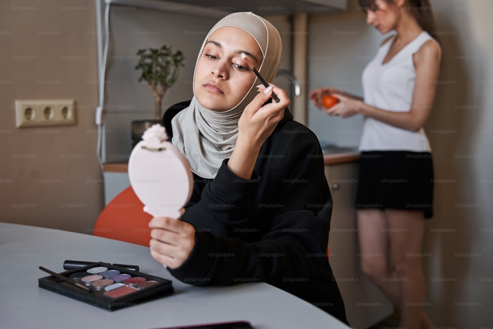 Porträtansicht der wunderschönen muslimischen Frau, die in den Handspiegel schaut und Schatten auf ihre Augenlider aufträgt, während ihre beste Freundin in der Küche Obst wäscht