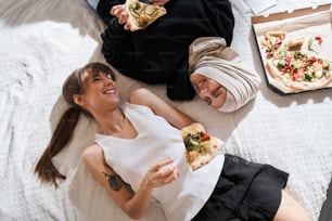 とても違います。ピザのスライスを持ってベッドに横たわり、喜びの笑顔でお互いを見つめている多様な学生の女の子の上面図。異なる信仰と友情のコンセプト