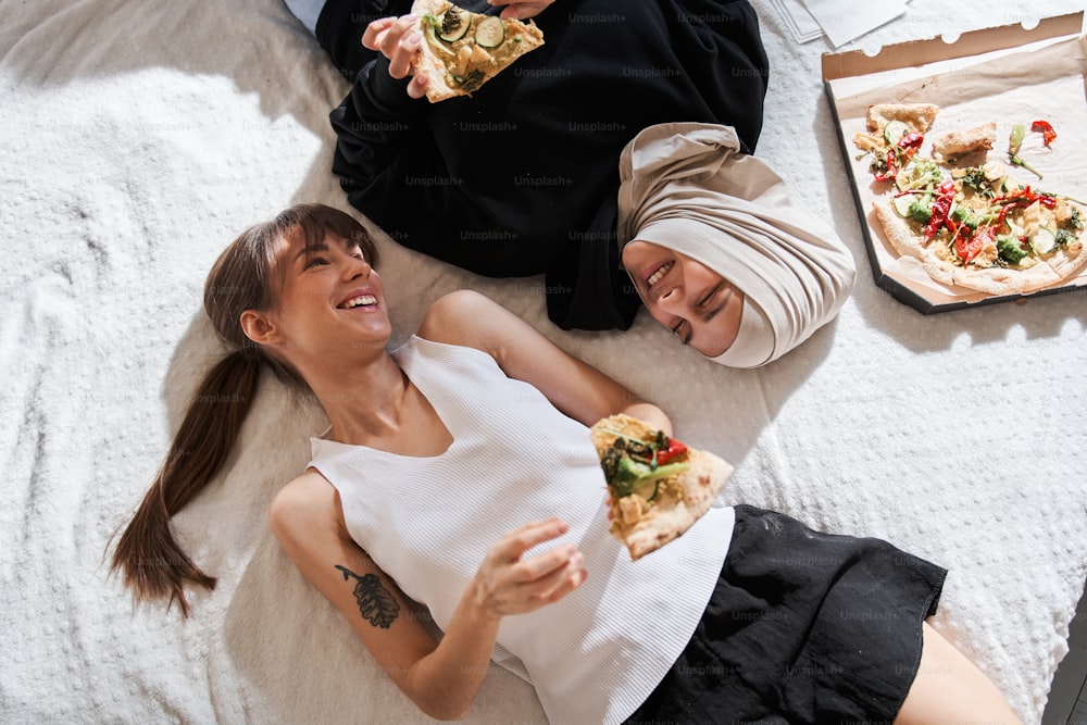 So different. Top view of the diverse student girls laying at the bed with slices of pizza and looking at each other with pleasure smile. Different faith and friendship concept