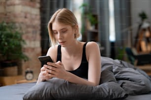 Mulher loira nova usando o telefone celular em seu trabalho on-line enquanto se senta na cama pela manhã