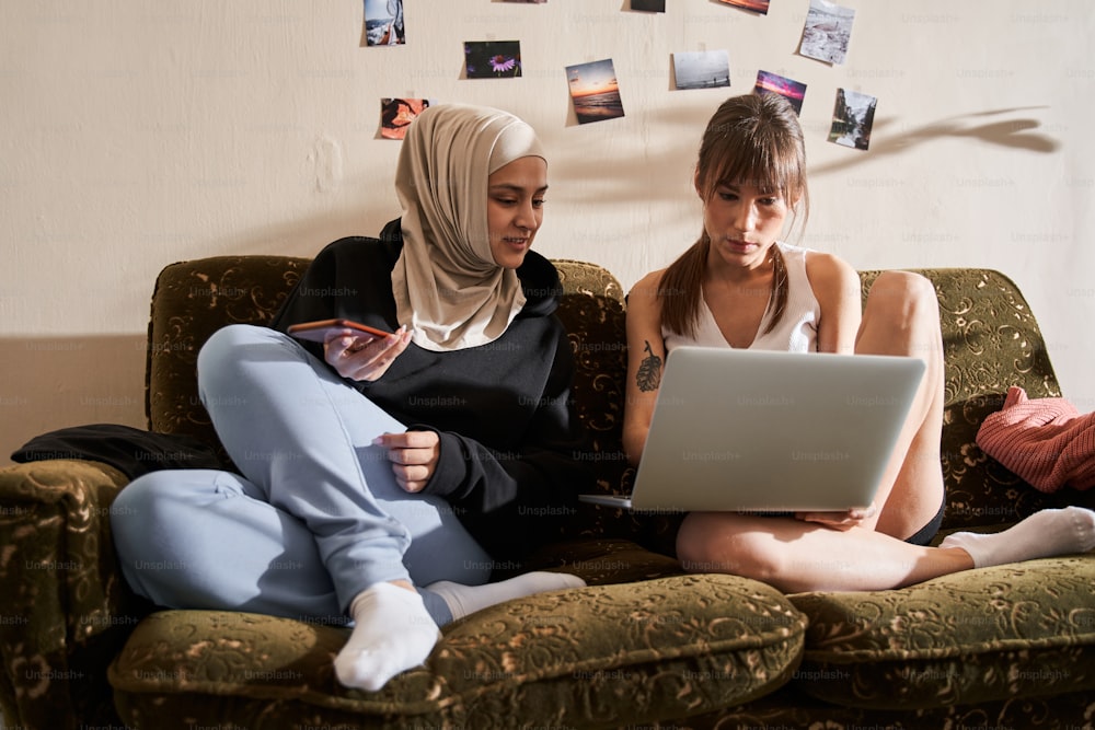 異なる信仰の2人のダイバー学生の女の子がソファに座って一緒に勉強しています。イスラム教徒の女性がノートパソコンの画面を覗き込み、ガールフレンドに何かについて尋ねます。ストックフォト