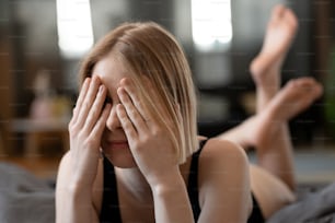 Joven rubia cubriéndose los ojos con las manos acostadas en la cama en el dormitorio