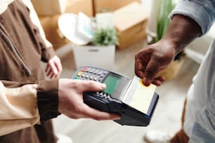 Hand eines jungen Mannes in Arbeitskleidung, der ein Zahlungsterminal hält, während ein afrikanischer Mann für das Laden und die Lieferung per Kreditkarte bezahlt