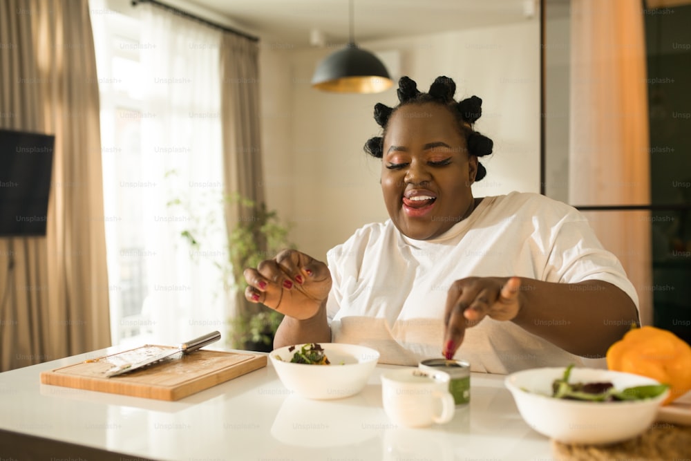 Schöne ruhige multirassische Frau, die ihr Gericht probiert und am Tisch um das Gemüse sitzt, während sie gesundes Essen in der Küche zu Hause kocht. Stockfotografie