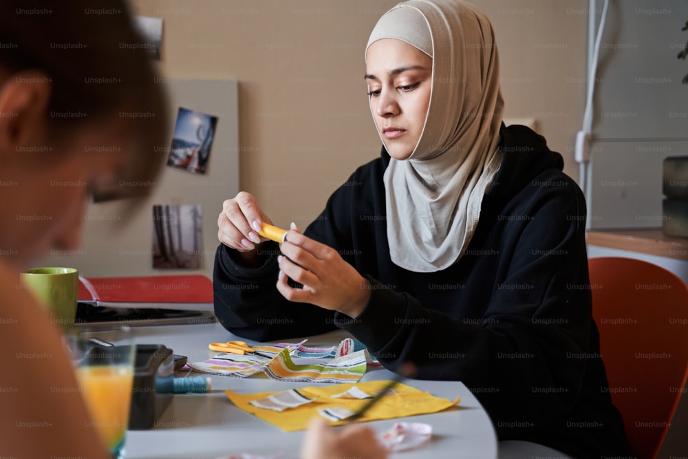 ヒジャーブを着たイスラム教徒の女性が糸のスプールを持ち、自宅で友人とテーブルで縫う腰のポートレートビュー。手工芸品と趣味のコンセプト