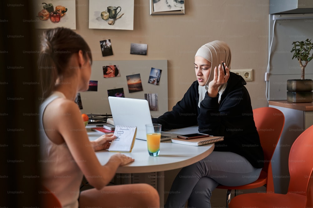 テーブルに座ってノートパソコンの画面を見て、グループメイトと一緒に試験の準備をしながら何かを言うイスラム教徒の学生の女の子の全身像。ストックフォト