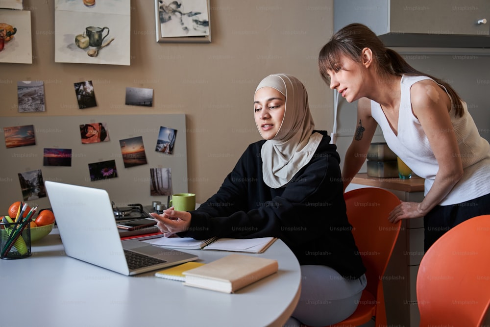 Vista a figura intera della donna multirazziale che indica lo schermo del laptop con il dito mentre consiglia qualcosa con il suo compagno di gruppo caucasico durante lo studio a casa