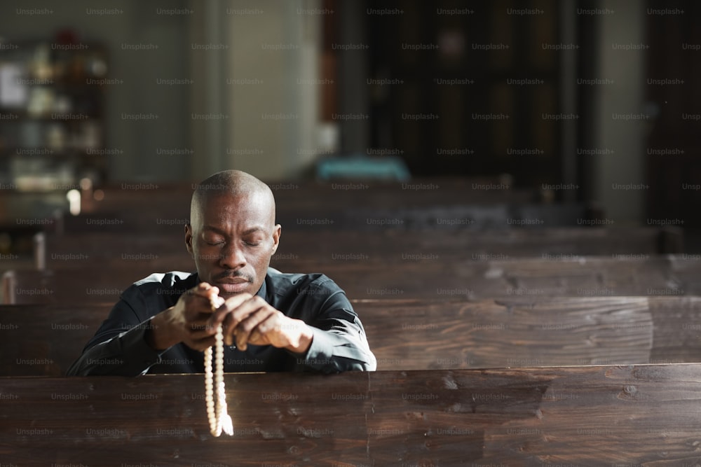 Afrikanischer Mann, der in der Kirche sitzt, Perlen hält und mit geschlossenen Augen betet