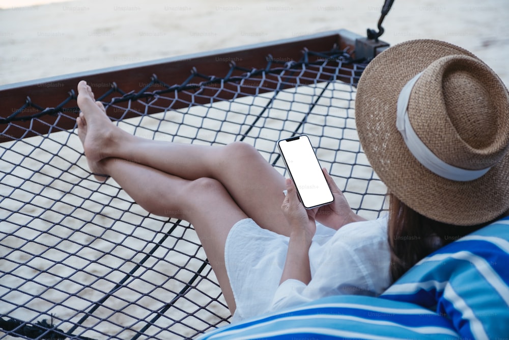 Image de maquette d’une femme tenant un téléphone portable avec un écran de bureau vide allongée sur un hamac sur la plage