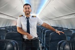 Positive confident pilot leaning on an empty passenger seat and smiling while standing in the aisle