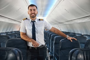 Piloto seguro de sí mismo apoyado en un asiento de pasajero vacío y sonriendo mientras está parado en el pasillo