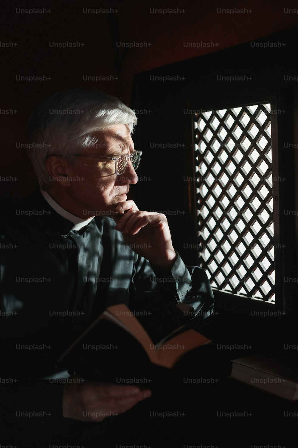 Serious senior priest sitting with Bible in confessional in the church