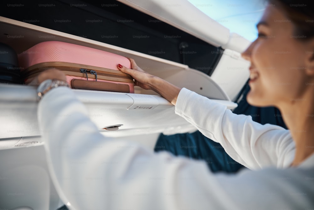 Cropped photo of a smiling young woman carefully putting her suitcase on a luggage shelf in a plane