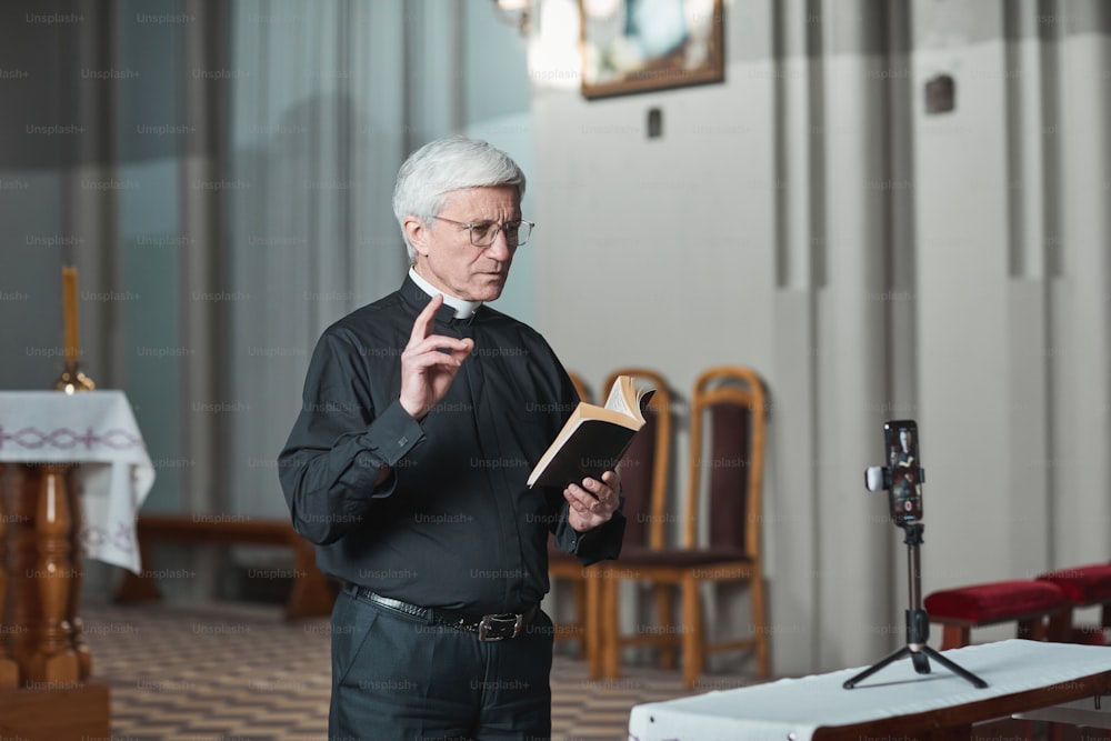 Sacerdote anziano che legge la Bibbia e riprende la sua performance con il cellulare mentre è in piedi in chiesa
