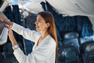 Cheerful young woman smiling while putting her carry-on luggage into the overhead compartment
