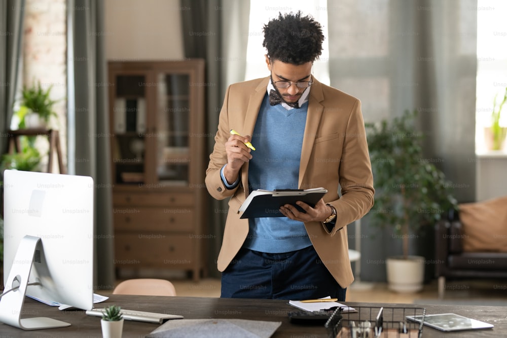Serious busy young black marketing manager in jacket and sweater standing at wooden table and working with papers in home office