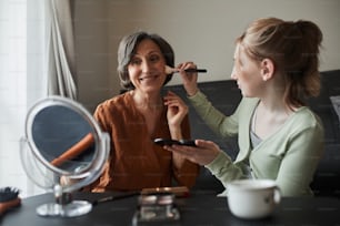Looking great. Happy senior woman looking at her reflection at the mirror with pleasure while her granddaughter preparing makeup for her at the cozy living room
