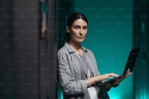 Waist up portrait of female data engineer holding laptop and looking at camera while working with supercomputer in server room, copy space