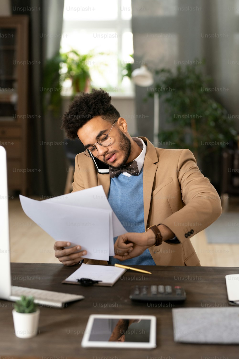 Serious multi-tasking young mixed race manager in bow tie sitting at wooden table with calculator and checking time while talking by phone and examining papers