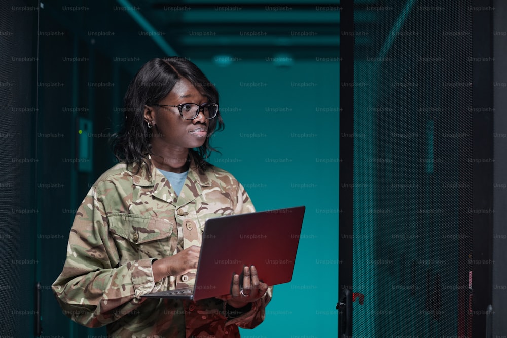 Retrato da cintura para cima da mulher afro-americana jovem usando o uniforme militar usando o laptop enquanto está na sala do servidor, espaço da cópia