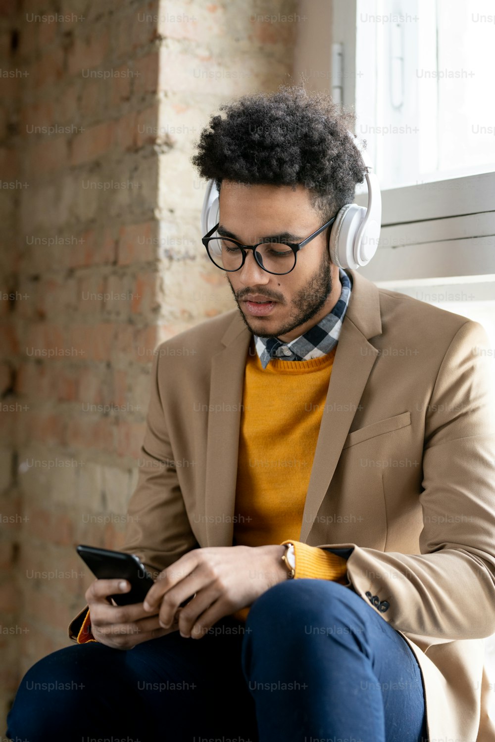 Modern young black businessman with afro hairstyle sitting on window sill and searching for track on smartphone while listening to music in wireless headphones