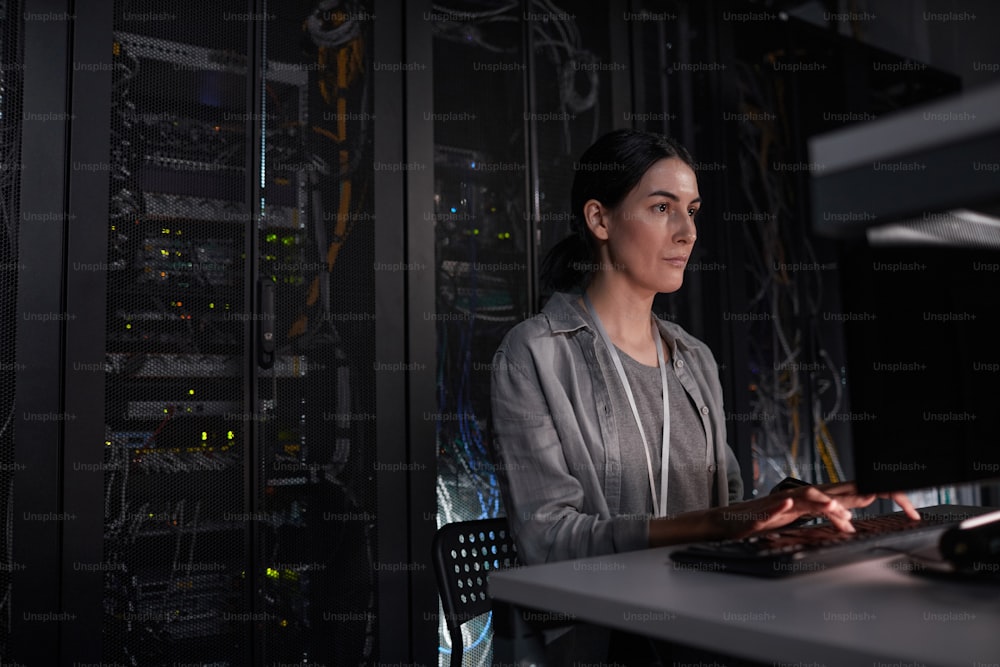 Portrait of female server engineer using laptop while sitting in dark IT room, copy space