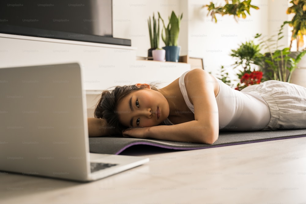 Before the training. Full length view of the asian woman laying at the mat at the floor and watching yoga lesson at the laptop before her training. Stock photo