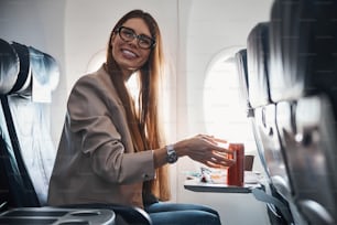 Female with long brown hair sitting at window seat and grabbing red can of fizzy drink while smiling for camera