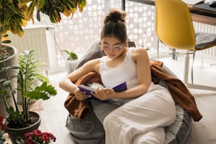 Ganzkörperansicht des jungen multirassischen Mädchens, das ein Buch hält, während es zu Hause liest oder lernt. Ruhige Millennial-Frau, die sich auf die Prüfungen vorbereitet oder ihr Wochenende verbringt