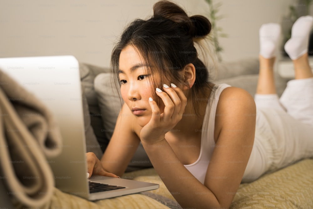 Portrait view of the pleasant asian brunette girl laying at big bed in modern room, looking at the screen with serious emotions. Girl surfing web, making purchases online or booking flight tickets