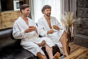 Two man in bathrobe sitting on sofa in living room of spa house and drink a tea. Relax and wellness concept. Spending and enjoy time together.