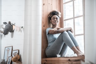 Sadness, emotion. Depressed young dark skinned woman barefoot in home clothes sitting on windowsill looking into room