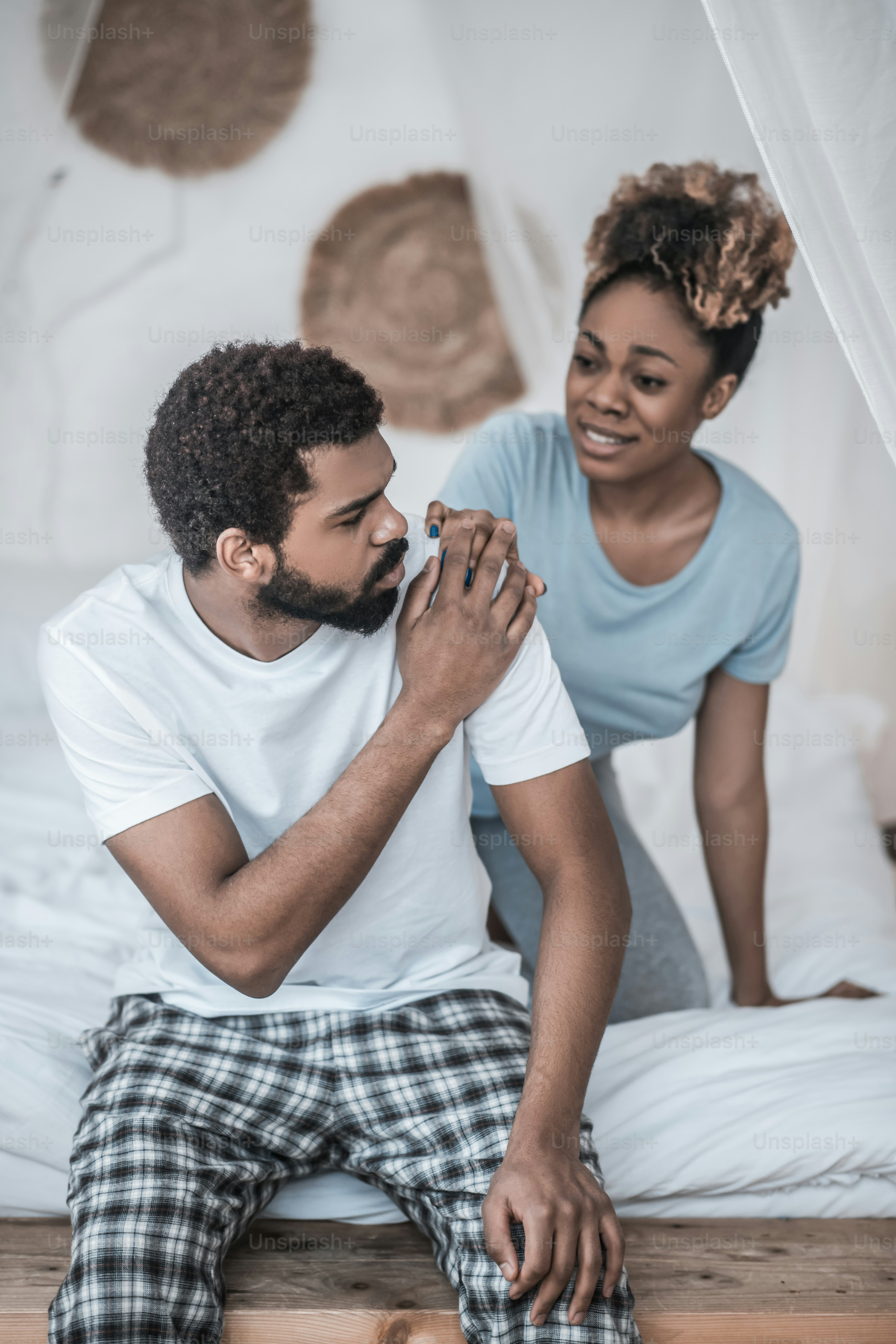 Reconciliation. Sad young dark skinned husband and anxious distressed wife touching his shoulder at home on bed