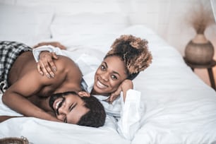 Morning, day off. Cute young adult african american woman in white shirt hugging laughing bearded husband lying on bed at home