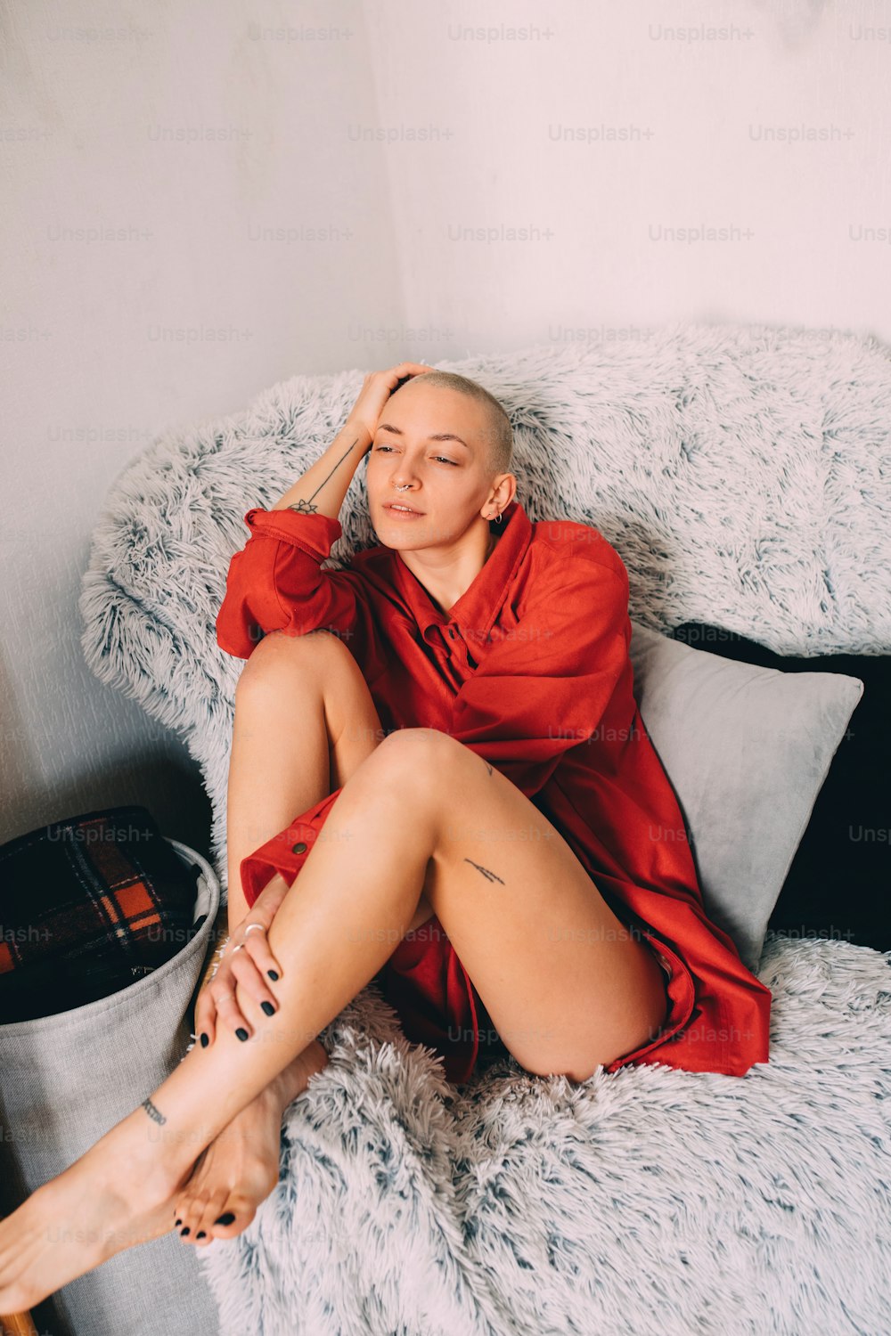 Full length view of the short haired tattooed woman wearing red shirt sitting at the armchair at the fur at home and thinking about something. Home lifestyle concept. Stock photo