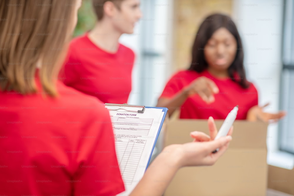 Formular, Spende. Freiwilliger im roten T-Shirt mit Dokument und Marker in den Händen, mit dem Rücken zur Kamera und Kollegen mit Kisten
