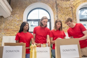Trabajo en Equipo. Estudiantes voluntarios serios comprometidos con camisetas rojas empacando cajas de donaciones amigablemente en el centro de caridad
