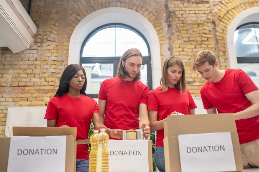 Gemeinschaftsarbeit. Engagierte ernsthafte studentische Freiwillige in roten T-Shirts, die Spendenboxen freundschaftlich im Wohltätigkeitszentrum packen