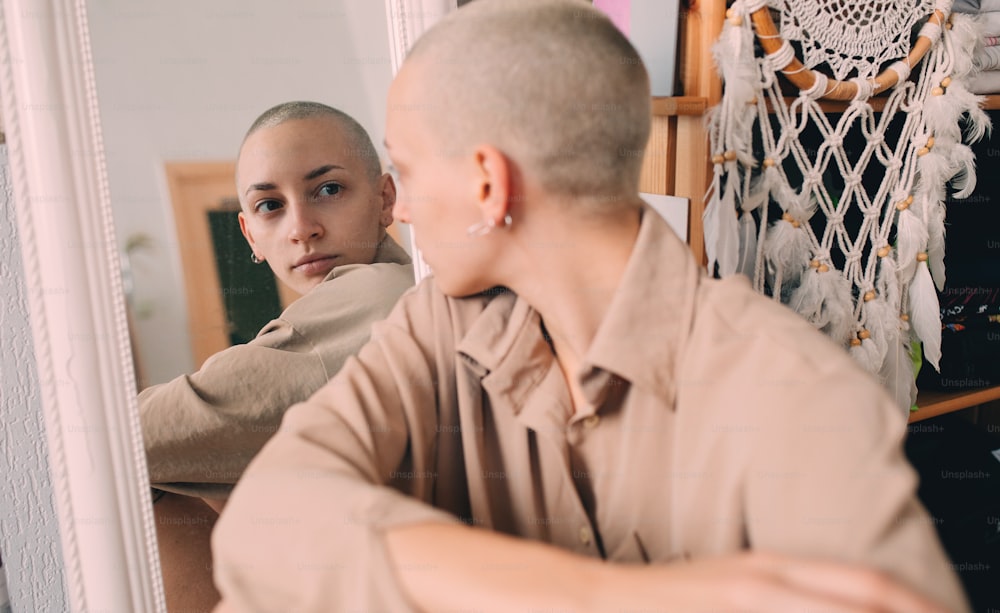 Waist up portrait view of the hipster girl with short hair and tattoos sitting at the floor and looking to the mirror at her reflection. Woman lifestyle concept. Stock photo