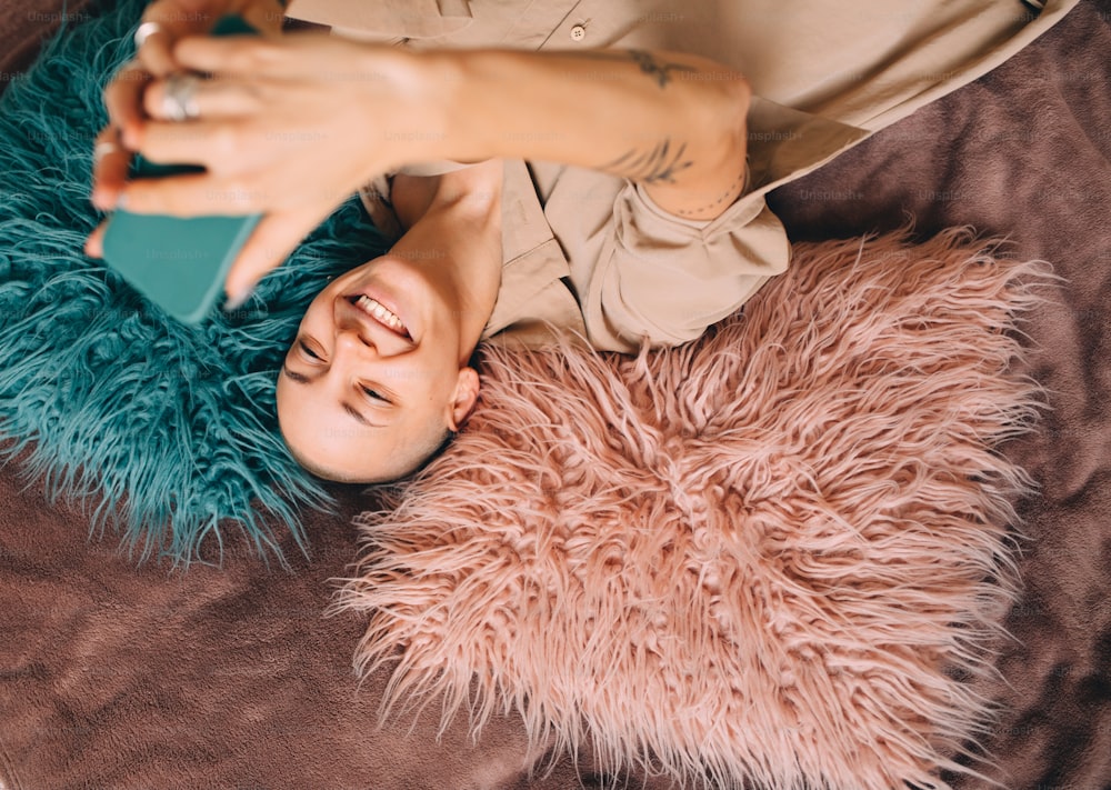 Taking selfie. Top view of the hipster tattooed girl with short haircut laying at the fur and posing to the selfie at her cell phone. Woman lifestyle and technologies concept. Stock photo