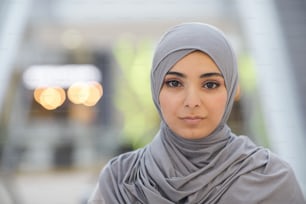 Close up portrait of beautiful Middle-Eastern woman wearing grey headscarf while standing in city with blurred background, copy space