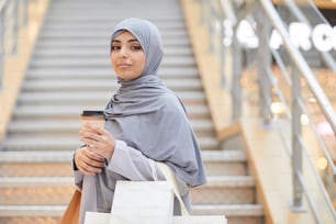 Retrato de cintura para arriba de una joven del Medio Oriente con pañuelo en la cabeza y sosteniendo una taza de café mientras disfruta de las compras en el centro comercial, copie el espacio