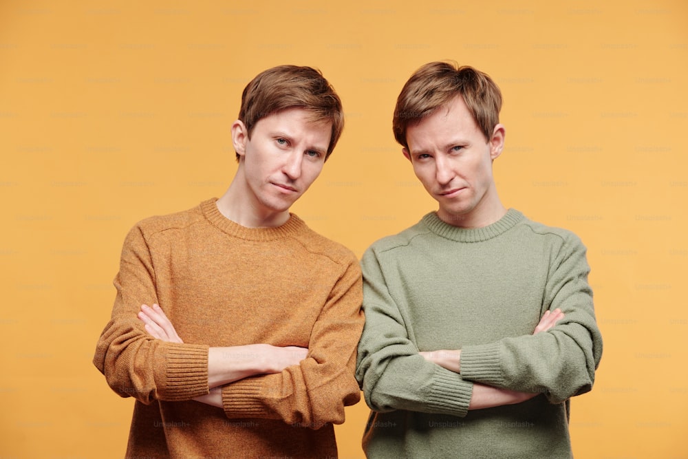 Portrait of frowning tween brothers in mustard and olive sweaters standing with crossed arms against orange background