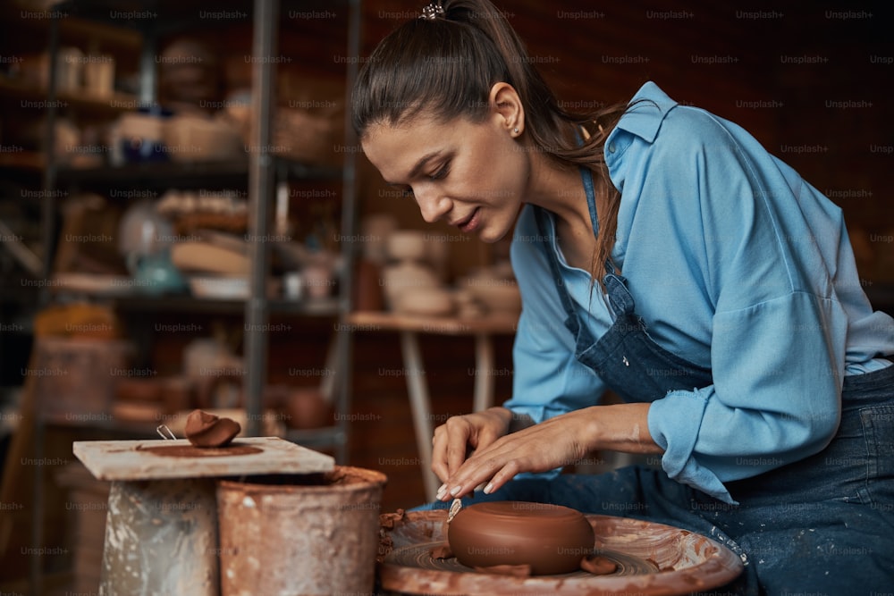 Hermosa artesana morena con delantal sentada en la rueda de cerámica mientras hace cerámica y vajilla de cerámica en el estudio de arte