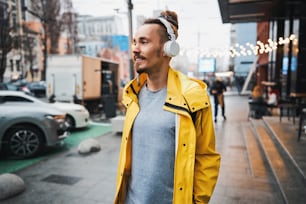 Happy handsome stylish guy with dredlocks is listening to music while going along urban street