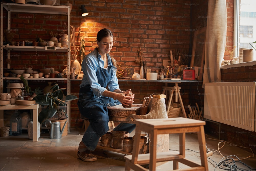 Retrato de vista lateral de comprimento total de bela artesã feminina focada sentada no estúdio de arte enquanto molda o produto cerâmico da argila fresca