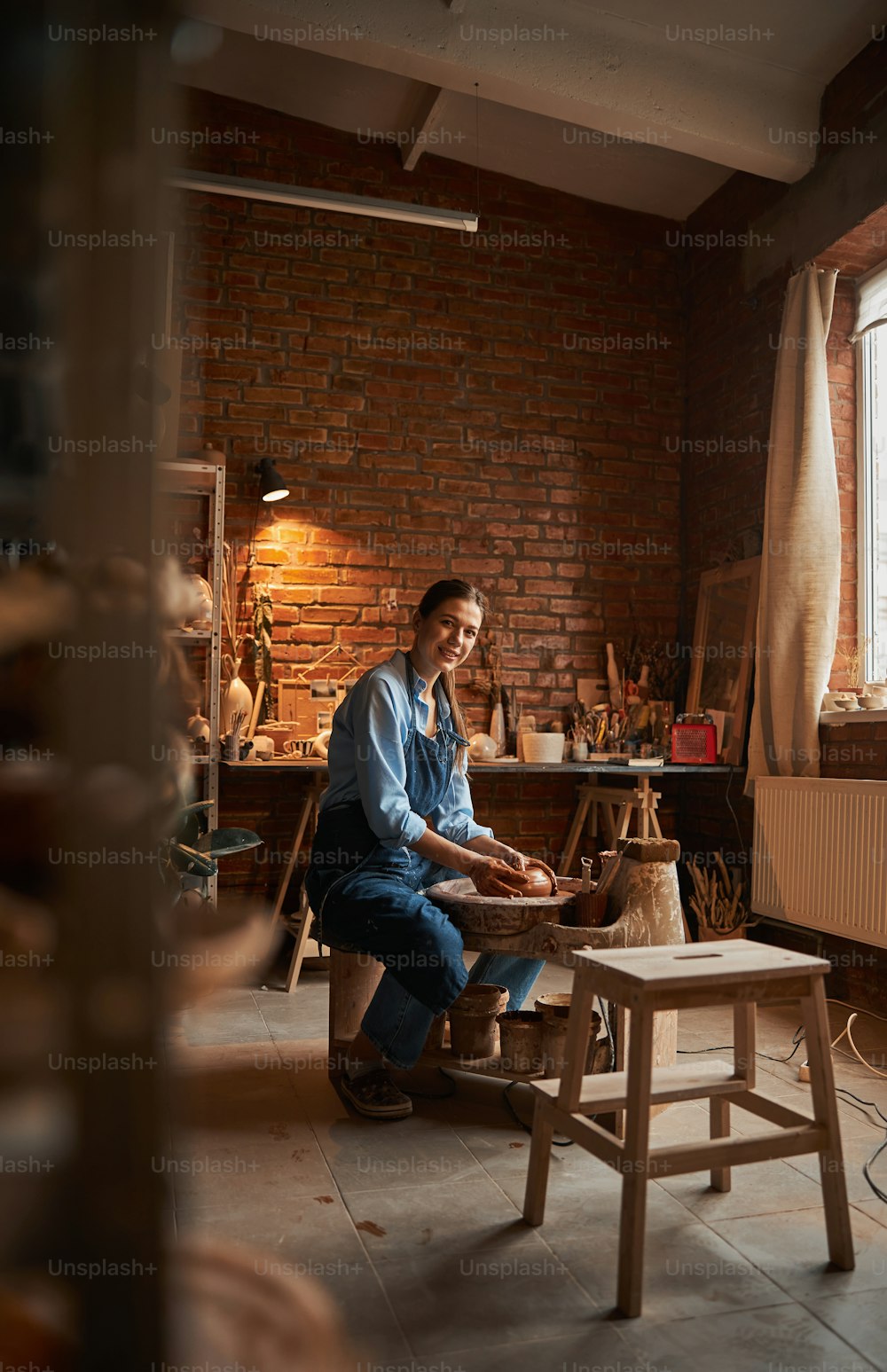 Retrato de vista lateral de cuerpo entero de una hermosa y elegante artesana sentada cerca de la rueda de cerámica mientras hace algo en el estudio de arte
