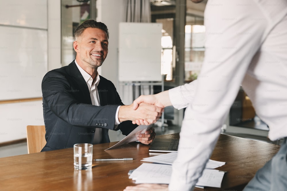Business, career and placement concept - satisfied director man 30s smiling and shaking hands with male candidate who was recruited during interview in office