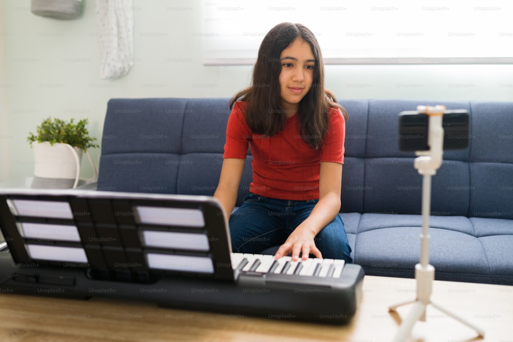 Prendre des cours de musique en ligne. Préadolescente latine prêtant attention à un appel vidéo avec un professeur de piano sur son smartphone alors qu’elle est assise sur son canapé avec un clavier