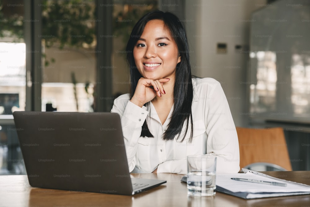 Image of beautiful asian business woman wearing white shirt looking at camera and smiling while sitting at table in office during work on laptop